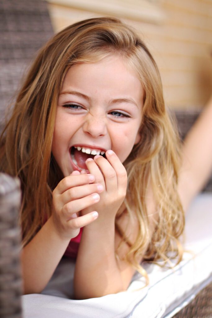Ein Mädchen hält ihre linke Hand leicht vor ihren lachenden Mund und zeigt doch ihre hellen Zähne.