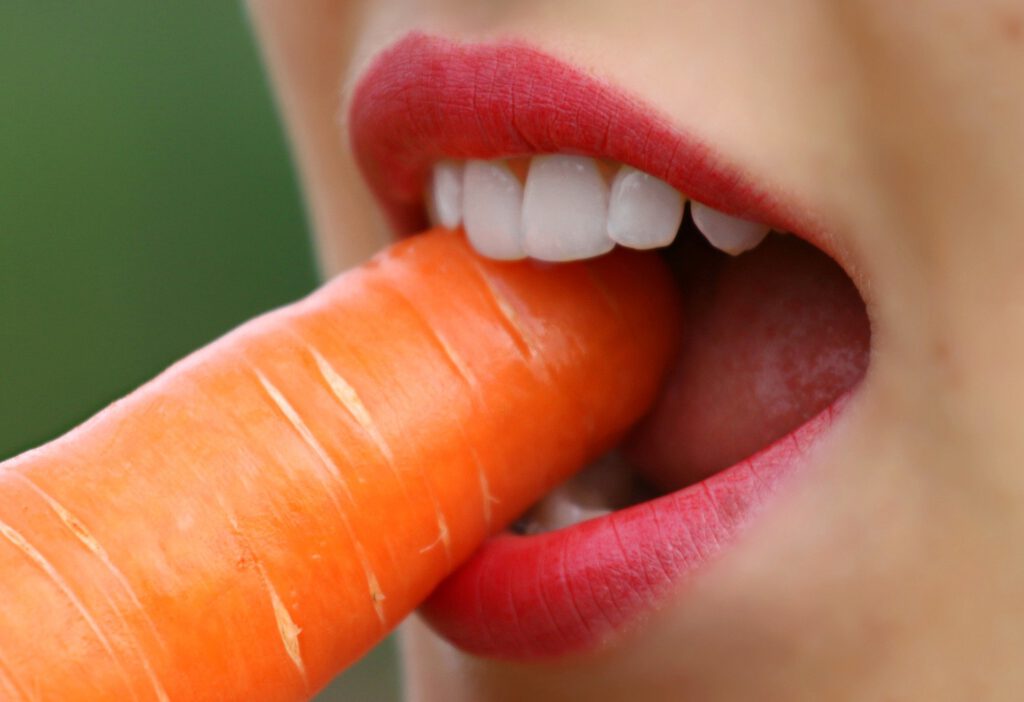 Ein Mädchen zeigt ihre knallroten Lippen und beißt mit ihren langen festen Zähnen auf eine Karotte, Zahnarzt, Zahnarztpraxis, Feste Zähne