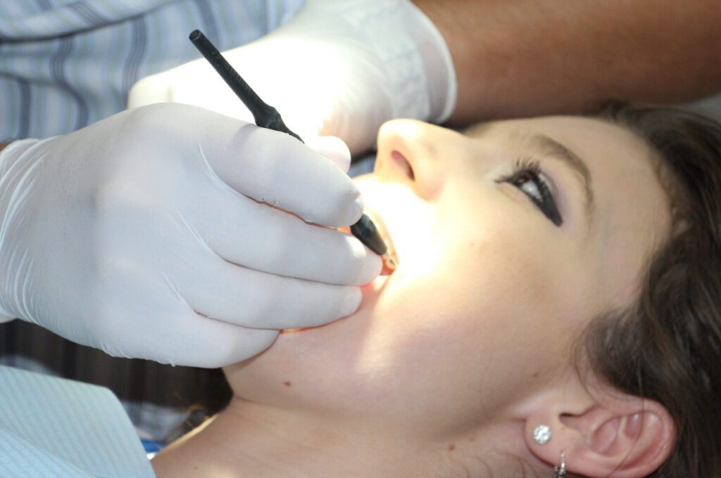 Eine dunkelhaarige Frau liegt auf dem Zahnarztstuhl. Man sieht nur ihren Kopf und eine Hand die in ihrem geöffneten Mund arbeitet.
