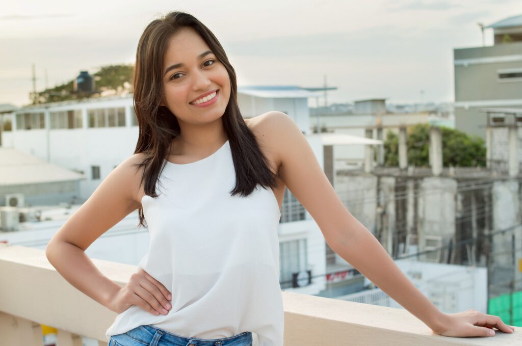 Eine junge Frau mit langem dunklen Haar steht mit ihrem weißen T-Shirt lächelnd auf einem Flachdach und lächelt dem Betrachter selbstbewusst zu. Knirscherschiene