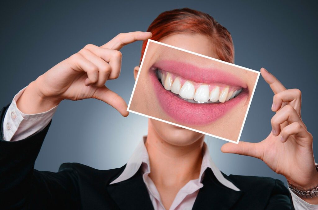 Eine dunkelblonde Frau hält mit fast gespreizten Fingern ein Poster mit schönen Zähnen vor ihr Gesicht, Implantate, Zahnimplantate