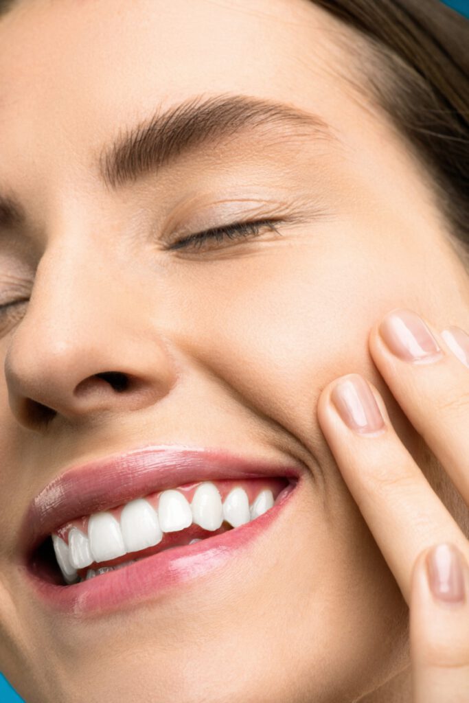 Eine Frau legt lächelnd ihre linke Hand auf ihre linke Wange, während sie ihre weißen Zähne umrandet von ihren roten Lippen zeigt, Implantate