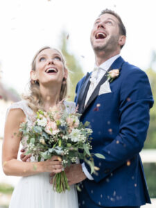 Ein glückliches Hochzeitpaar blickt mit Staunen und offenem Mund in den Himmel, sie trägt ein weisses Kleid und hält den Hochzeitsstrauß vor sich er trägt einen dunkelblauen Anzug